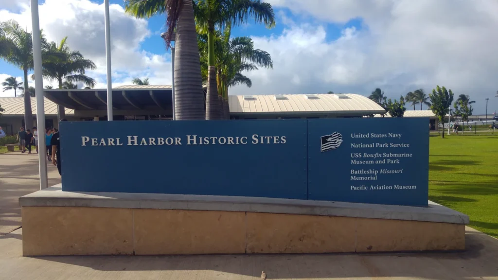 שלט כחול של Pearl Harbor Historic Sites בכניסה לאתר, עם עצי דקל ושמיים מעוננים באואהו